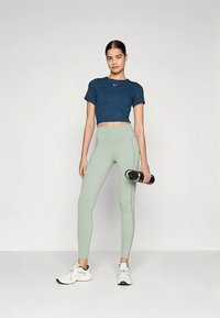 O conjunto desportivo inclui um top curto azul escuro e leggings de cintura alta verde-claro. A modelo segura uma garrafa de água preta e usa sapatilhas brancas.