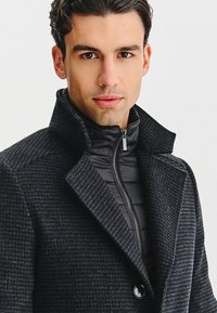 Czarny płaszcz wełniany z mieszanki włókien o teksturowanym wykończeniu, z wysokim kołnierzem i czarną ocieplaną wewnętrzną kurtką zapinaną na zamek.