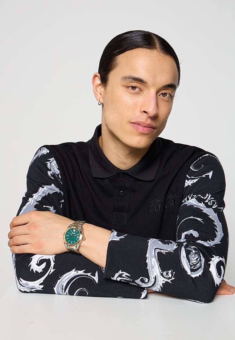 Polo noir à texture avec des motifs blancs élaborés sur les manches. Modèle portant une montre dorée avec un cadran vert, les bras posés sur une surface plane.