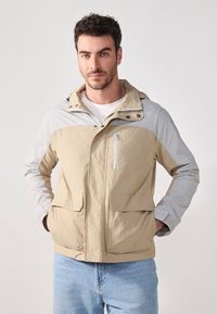 Beige og grå hettejakke med glidelås-flest lomme, to frontlommer og elastiske mansjetter. Laget av lettvektsstoff med en jevn tekstur.