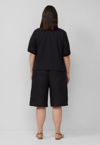 Schwarzes Kurzarmoberteil mit gerafften Ärmeln, kombiniert mit lockeren schwarzen Shorts. Beides aus leichtem Stoff gefertigt. Sandalen runden den Look ab.