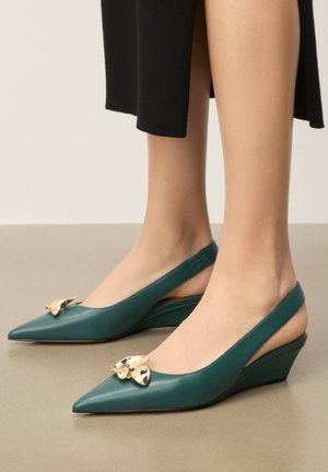 Teal leather slingback wedge heels with pointed toes and gold butterfly embellishments worn with a black skirt.