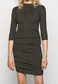 Svart, kroppsnära klänning med en texturerad, metallisk stickning. Har trekvartslånga ärmar och draperade tygdetaljer runt midjan.