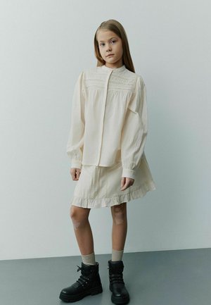 Blusa beige chiaro con dettagli testurizzati, abbinata a una gonna svasata coordinata. Stivaletti neri alla caviglia con lacci completano l'outfit.