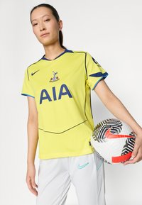 Žuta Nike nogometna majica s plavim detaljima i "AIA" logom, uparena sa sivim hlačama. Drži šareni nogomet s crvenim Nike logom.