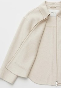 Beige wool blend jacket featuring a zip closure, stand collar, and side pockets with a smooth texture and slightly curved hem.
