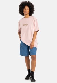 Lyserød oversized T-shirt med et cirkulært grafisk design på brystet, parret med blå denim shorts og sorte træningssko.
