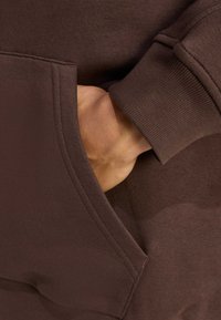 Sudadera marrón con una tela texturizada, con un bolsillo tipo canguro y puños acanalados; una mano parcialmente metida en el bolsillo.