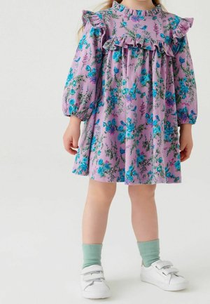 Enfant portant une robe florale violette avec un devant à volants et des manches longues bouffantes, des chaussettes vert clair et des baskets blanches à scratch.