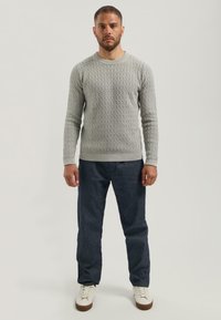 Man staat rechtop met het gezicht naar voren, gekleed in een lichtgrijze gebreide trui met textuur, donkerblauwe jeans en witte sneakers met bruine zolen.