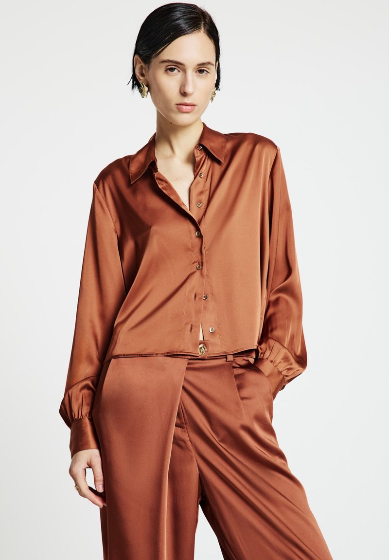 Woman with short dark hair wearing rust-colored satin button-up blouse and matching wide-leg trousers against plain background.