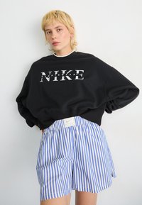 Crna dukserica s bijelim natpisom "NIKE", u kombinaciji s plavim i bijelim prugastim kratkim hlačama s elastičnim pojasom i oznakom loga.