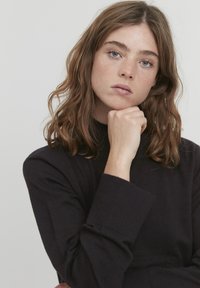 Pulover negru tricotat cu un guler înalt, mâneci largi și un finisaj texturat. Modelul pozează cu bărbia sprijinită în palmă, pe un fundal neutru.