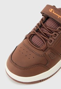 Adidași maro cu un upper din plasă texturată, închidere cu șireturi și curea, cutie perforată pentru degete și o talpă medie albă contrastantă.