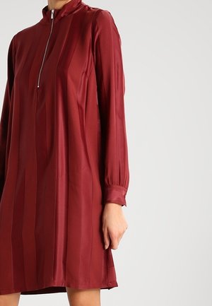 Bordeauxfarbenes Kleid aus glattem, glänzendem Stoff mit hohem Kragen, langen Ärmeln und einem halben Reißverschluss. Mit vertikalen Streifen.