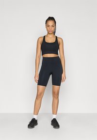 Musta urheiluliivit ja siihen sopivat korkeavyötäröiset shortsit joustavasta kankaasta. Malli seisoo yksiväristä taustaa vasten ja on pukeutunut mustiin lenkkareihin.