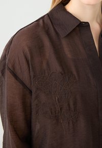 Camicia leggera marrone con colletto, caratterizzata da un ricamo intricato di un motivo floreale sul petto. Texture morbida con una finitura leggermente stropicciata.