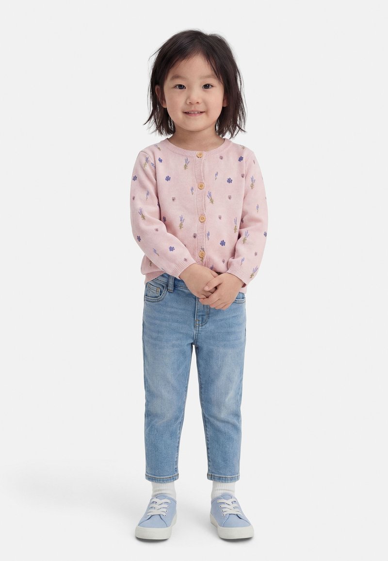 Νεαρό παιδί που στέκεται με τα χέρια σφιγμένα, φοράει ροζ πουλόβερ με λουλουδένιο σχέδιο, ανοιχτό μπλε τζιν, λευκές κάλτσες και μπλε αθλητικά παπούτσια, μπροστά σε λευκό φόντο.