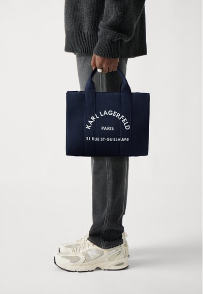 Тотебаг в тъмносин цвят, изработена от плат, с бял текст: "KARL LAGERFELD PARIS 21 RUE ST-GUILLAUME," и две здрави дръжки.