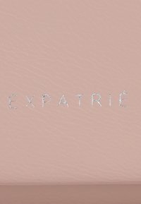 Vaaleanpunainen nahka tausta, jossa on kohokuvioitu teksti "EXPATRIÉ" hopeisena. Pintakuvio on sileä, jossa on hieno syy.