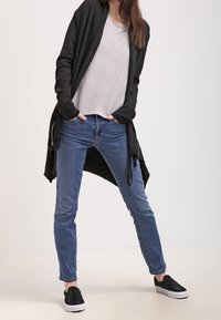 Černý cardigan přes šedé pruhované tričko, spárovaný s lehce modrými džíny a černými slip-on botami. Cardigan má volný střih a dlouhé rukávy.