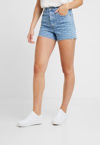 Denimshorts i ljusblått med ett mönster av "Levi's"-logotypen över hela ytan, fransat fåll och standard femficksdesign. Bärs med vita sneakers.