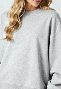 Vaaleanharmaa collegepaita, joka on valmistettu pehmeästä kankaasta. Paita on varustettu pyöreällä kaula-aukolla ja lasketuilla hartioilla, ja siinä on resorit hihansuissa ja helmassa.