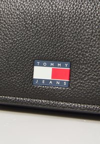 Svart teksturert skinnlommebok med en rektangulær blå og rød logo-patch med "TOMMY JEANS"-tekst og horisontalt fargeblokkmønster.