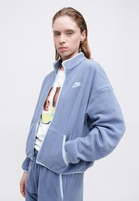 Svetlomodrá flísová bunda s vysokým golierom, zipsovým uzáverom, dvoma prednými vreckami a logom Nike na ľavom prsníku. Zladené svetlé akcenty.