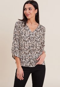 Camicia bianca con un motivo di foglie nere e arancioni, design con scollo a V, maniche a tre quarti e vestibilità rilassata. Pantaloni neri indossati sotto.