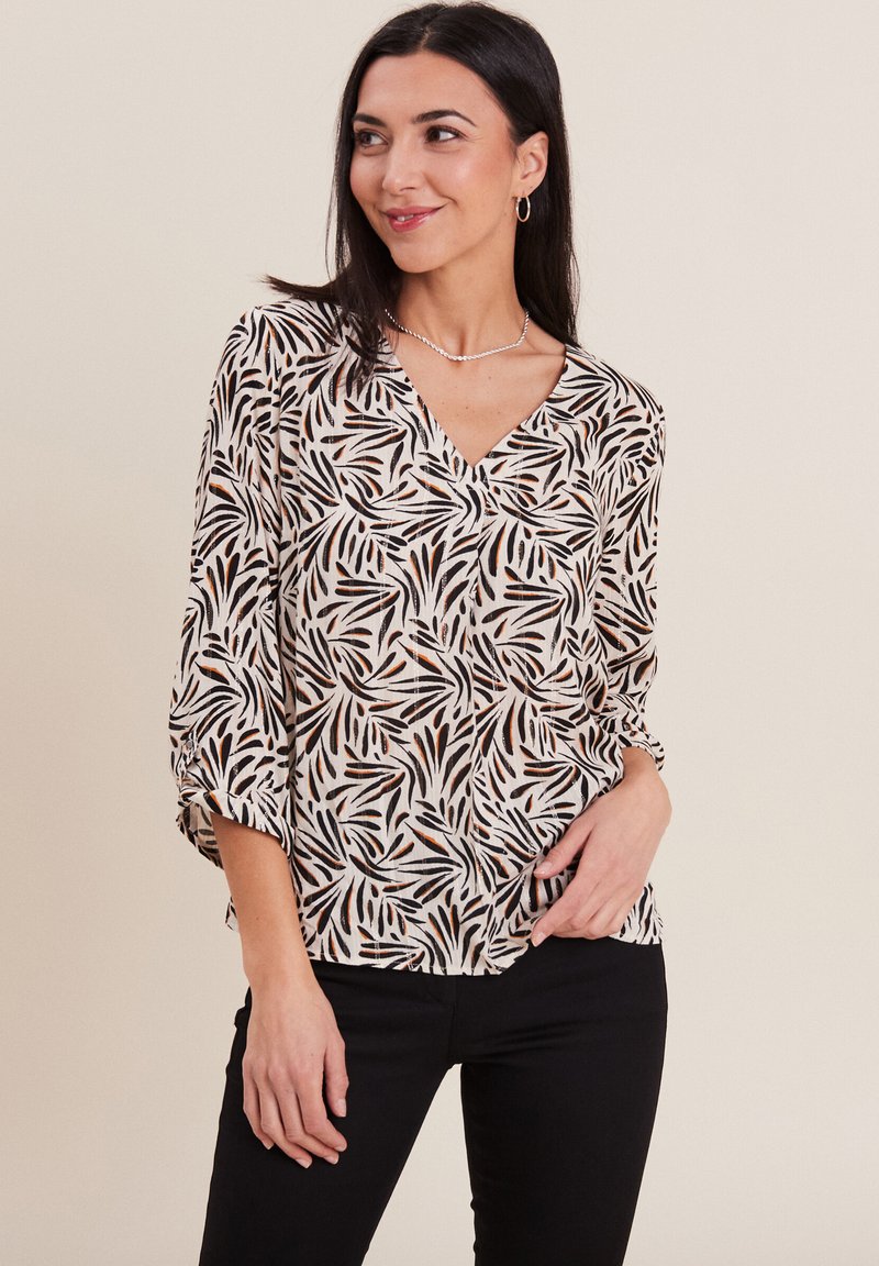 Camicia bianca con un motivo di foglie nere e arancioni, design con scollo a V, maniche a tre quarti e vestibilità rilassata. Pantaloni neri indossati sotto.
