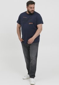 Mørkeblå kortermet t-skjorte med "Authentic Traditional Vintage" trykk, kombinert med mørkegrå jeans og hvite joggesko.