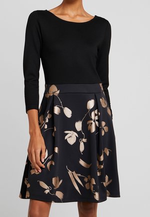 Kvinde iført en sort langærmet top kombineret med en knælængde navy nederdel med beige blomstermønstre.