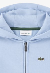 Light blue zip-up hoodie with green inner collar, black zipper, and small embroidered green crocodile logo on the left chest. Soft fabric.