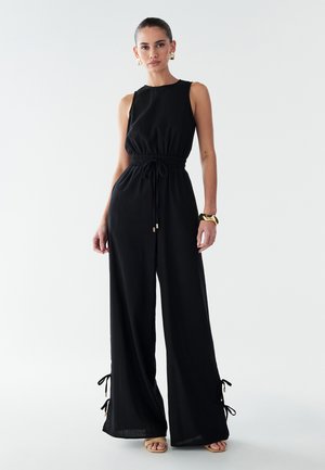 Black jumpsuit with a relaxed silhouette, elastic waistband, and adjustable ties at the cuffs. Smooth fabric with a slight sheen.