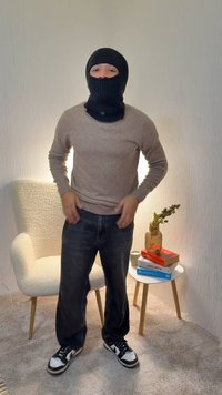 Crna pletena kapuljača, bež džemper, tamne traperice i crne tenisice s bijelim detaljima, stojeći u dobro osvijetljenom unutarnjem prostoru.