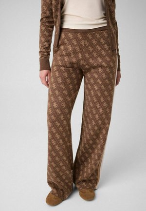 Personne portant un pantalon large marron à motifs et une veste assortie avec un haut beige et des chaussures marron, debout devant un fond uni.