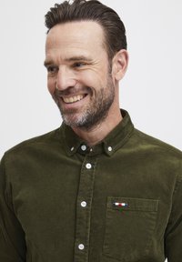 Mørkegrønn kordfløyelskjorte med knapp-ned krage, hvite knapper og en lommepatch med en liten rød, hvit og blå detalj.