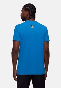 Niebieska koszulka typu T-shirt z krótkim rękawem, wykonana z gładkiego, syntetycznego materiału. Posiada czarną naszywkę z logo marki na górnej części pleców. Prosty design, dopasowany krój.