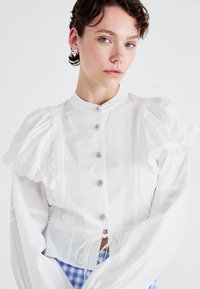 Hvit bluse med volangermuffer, blondekant, blå steinknapper og snøring i livet. Har høy krage og gjennomsiktige detaljer.