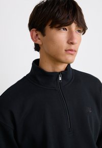 Sudadera de forro polar negra con cremallera y cuello alto, detalles en el hombro y un logo texturizado en el lado izquierdo del pecho. Tela suave.