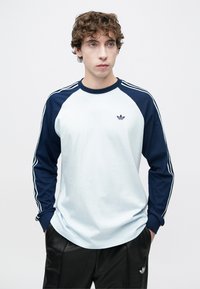 Μπλούζα με μακριά μανίκια, ανοιχτό μπλε σώμα και σκουρό μπλε μανίκια ράγκλαν. Διαθέτει τρεις λευκές ρίγες και ένα μικρό λογότυπο στο στήθος.