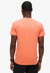 T-shirt a maniche corte di colore corallo arancione, scollo rotondo, materiale misto cotone, texture liscia e un piccolo etichetta sul lato sinistro in basso.