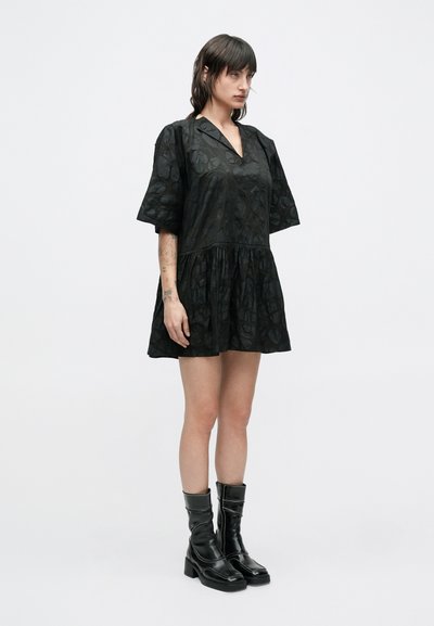 Μαύρο φόρεμα με κοντά μανίκια και υφασμάτινο μοτίβο, με V-λαιμόκοψη και ζώνη στη μέση. Συνδυάζεται με μαύρες μπότες μέχρι το γόνατο με ελαφρύ τακούνι.