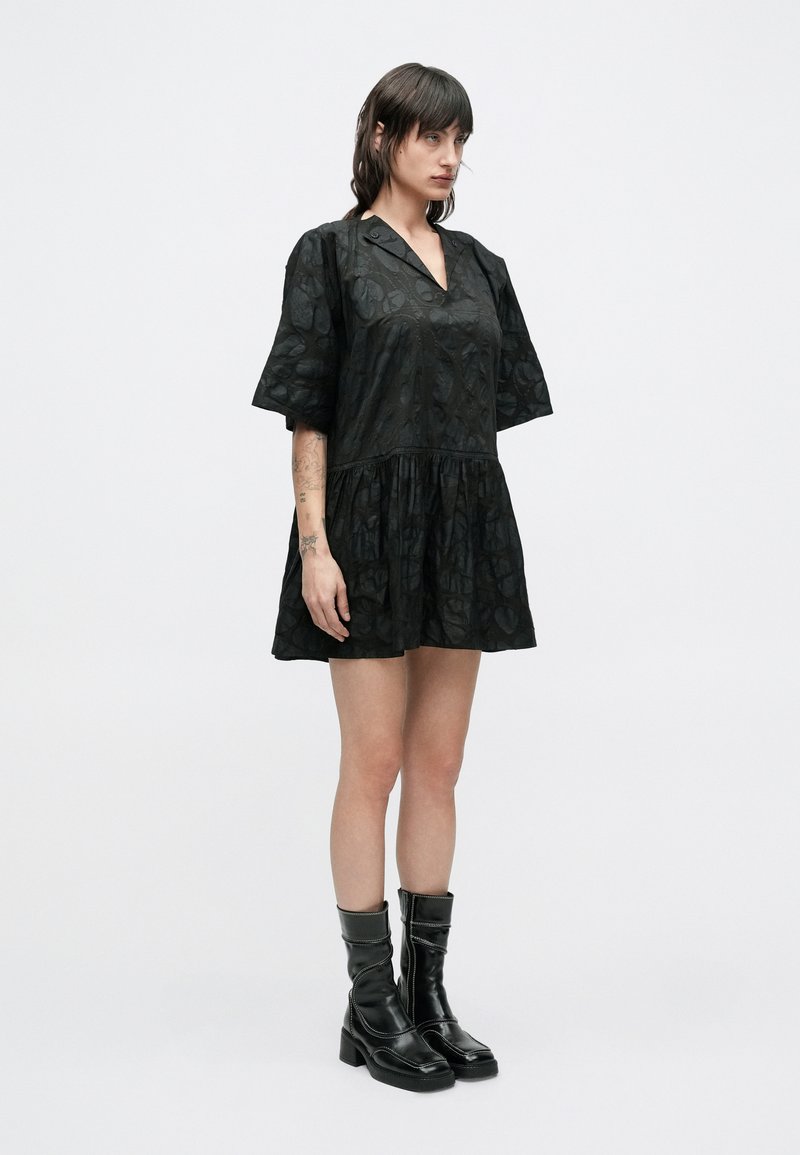 Robe noire à manches courtes avec un motif texturé, col en V et taille froncée. Associée à des bottes noires montant jusqu'au genou avec un léger talon.