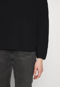 Czarny dzianinowy sweter o fakturowanej powierzchni, charakteryzujący się luźnym krojem i obniżonymi ramionami, w połączeniu z ciemnoszarymi dżinsami oraz widoczną ręką na dole.