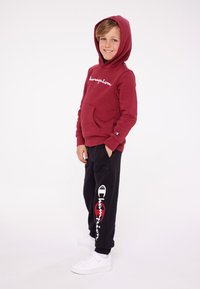 Felpa bordeaux con ricamo bianco "Champion", tasca frontale; pantaloni neri con logo rosso e testo bianco; sneakers bianche.