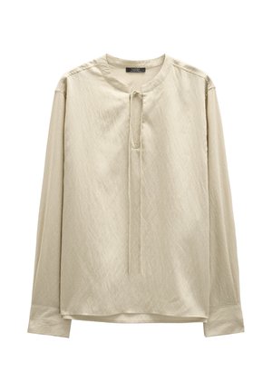 Koszula w jasnym beżowym materiale o teksturowanym wykończeniu, z okrągłym dekoltem i centralnym wiązaniem. Długie rękawy o luźnym kroju.