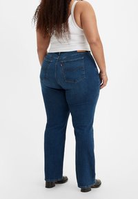 Kvinna med långt lockigt hår iförd mörkblå jeans, vit ärmlös topp och svarta klackstövlar, stående med ryggen vänd.