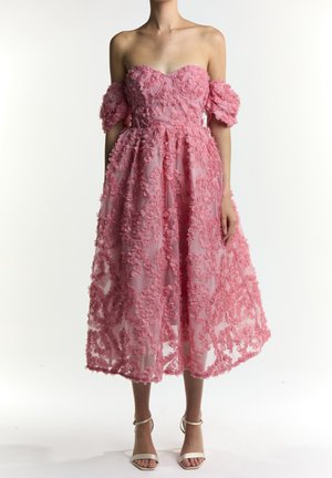 Różowa sukienka midi bez ramiączek z kwiatowym aplikacją 3D, z opuszczonymi bufkami na ramionach, noszona przez modelkę stojącą w białych szpilkach z paskiem wokół kostki.